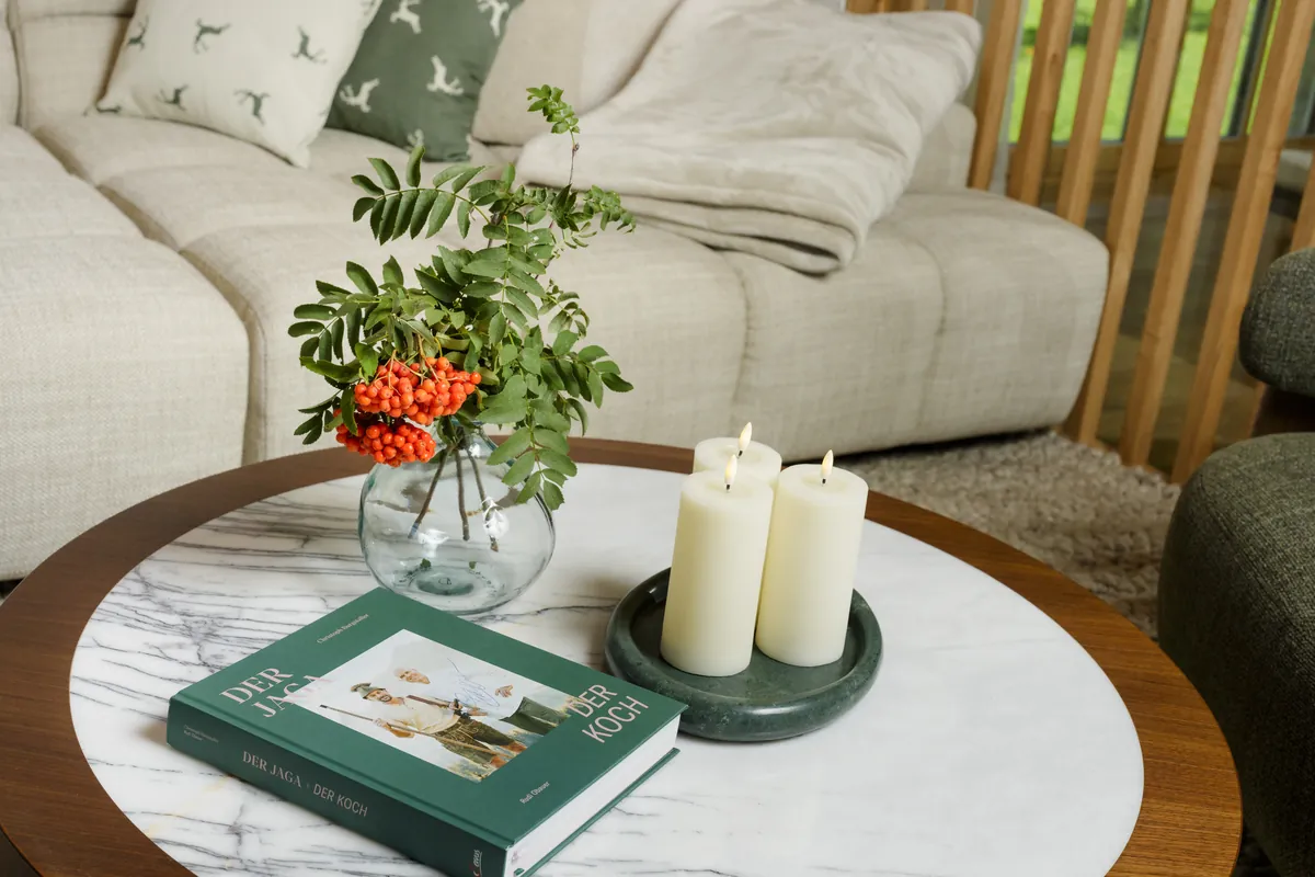 Runder Marmor-Couchtisch mit Kerzen, Vase mit Vogelbeerzweig und grünem Buch vor einem hellen Sofa im Chalet Gams